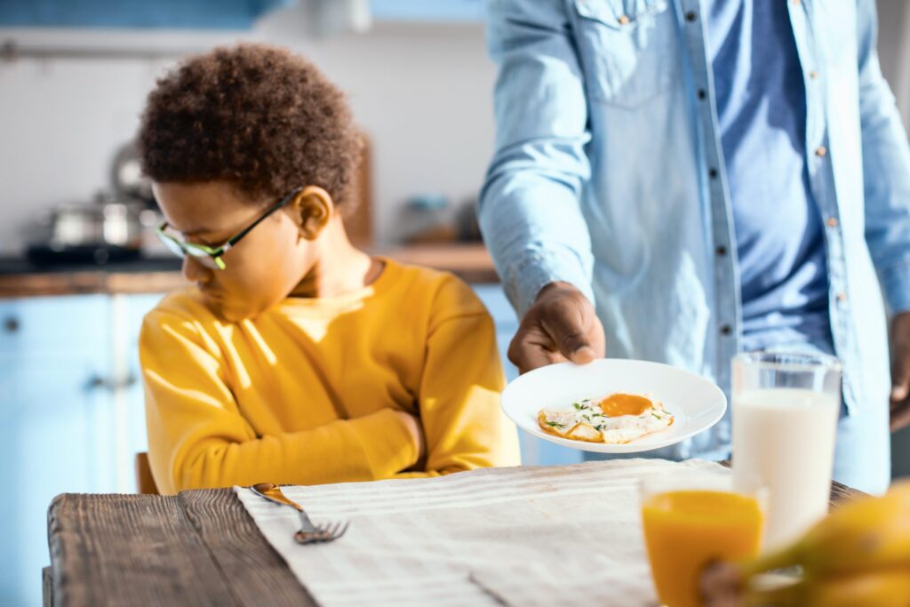 A child in a yellow shirt sits at a table with folded arms, looking away. An adult in a blue shirt offers a plate with eggs—perfect food for extremely picky eaters. The table has a glass of milk, a glass of orange juice, and a fork on a napkin.