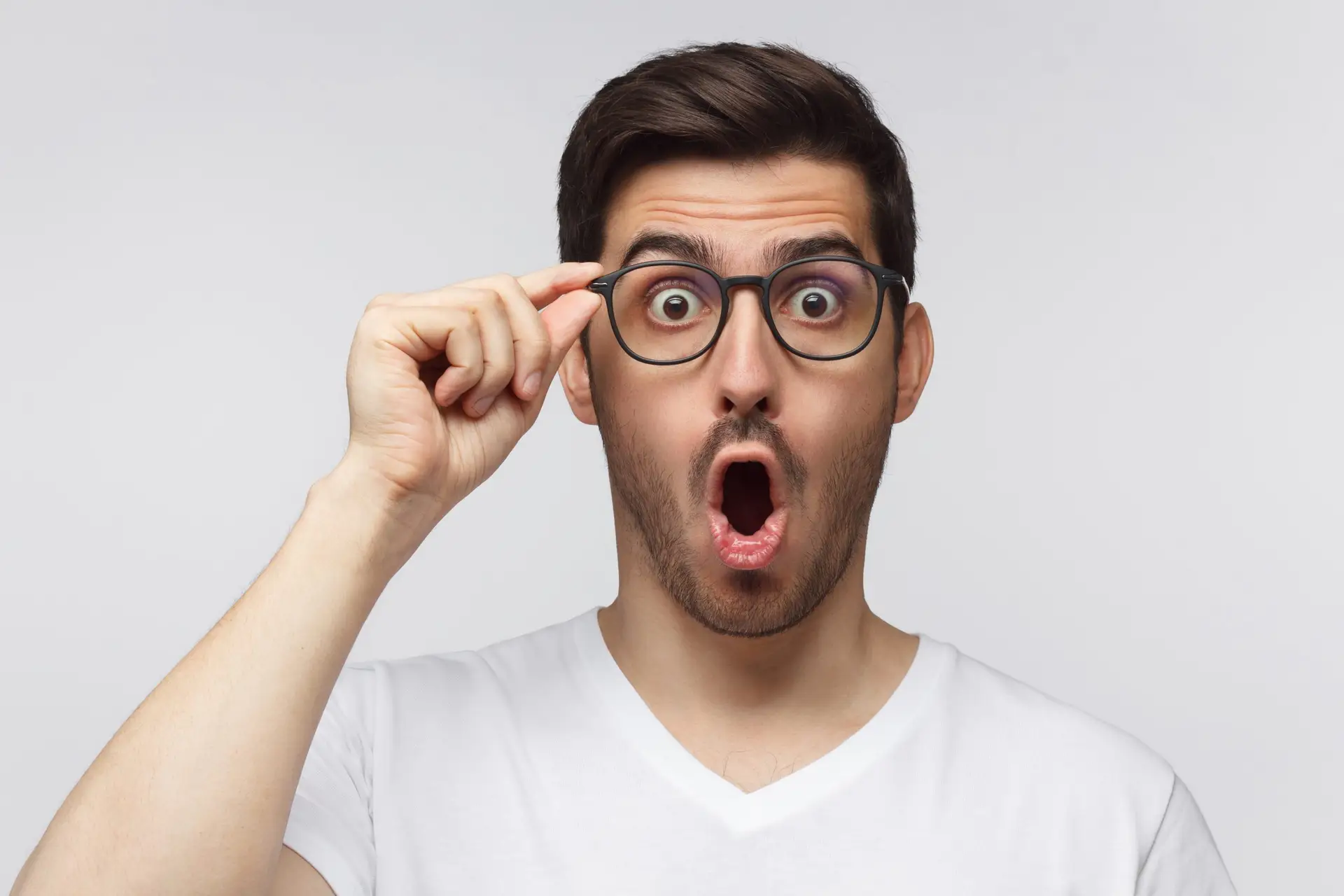 A surprised man with dark hair and glasses, embodying a sophisticated strategy for marketing, wears a white t-shirt. His mouth is open in disbelief as he holds his glasses with one hand against a plain white background, capturing the essence of advertising through outrage.