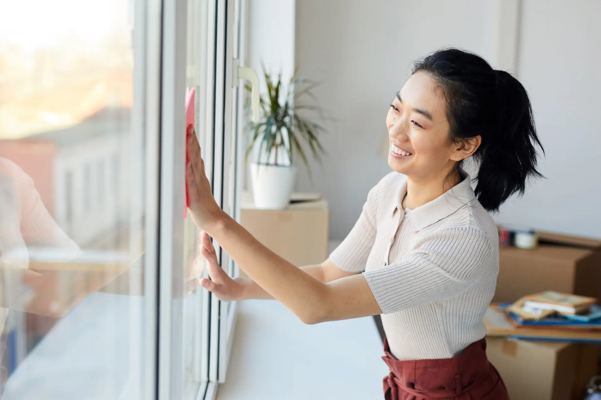 A woman smiles while engaging in some spring cleaning, using a pink cloth to clean a large window in a bright room. A potted plant and a box are visible in the background, reminding us of the importance of workspace tidiness and safety tips.