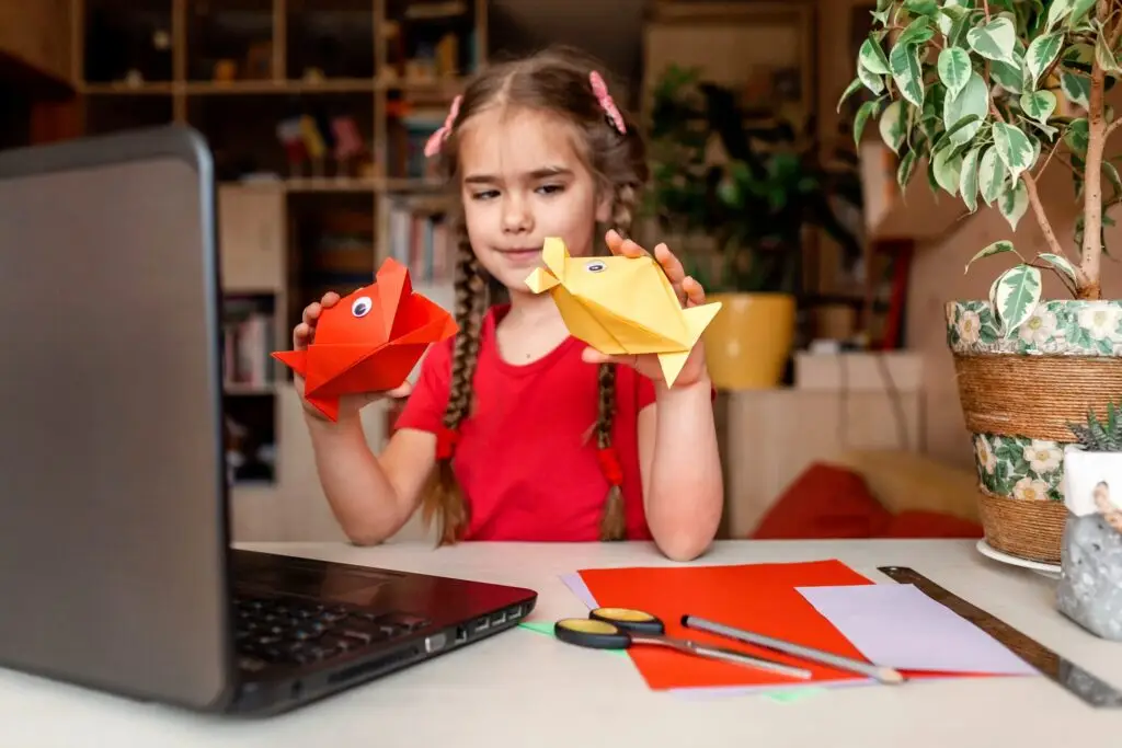 A young girl with braided hair, donning a red shirt, sits at a table holding two origami fish—one red and one yellow. She gazes at a laptop displaying "Three Easy Origami Paper Crafts: Step by Step," with paper and scissors scattered on the table. A plant adds greenery in the background.