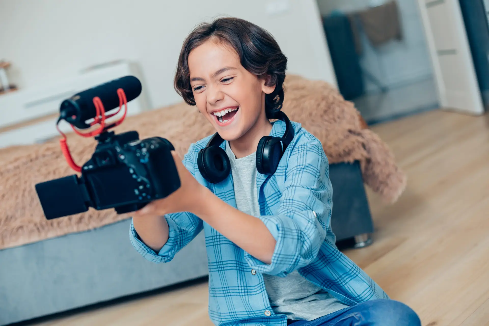 A smiling child in a blue plaid shirt and headphones holds a camera with a microphone attached, ready to share their top ten tips for kids starting a YouTube channel. They are sitting on the floor in a cozy room with a bed in the background.