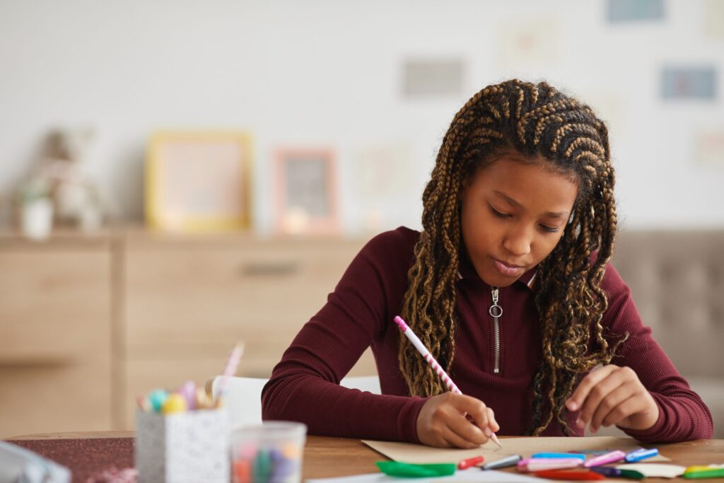 A young girl with braided hair, demonstrating fine motor skills, sits at a table focused on writing with a pencil. The table is scattered with colorful markers and pens. She wears a maroon top, and the background shows blurred frames on the wall.