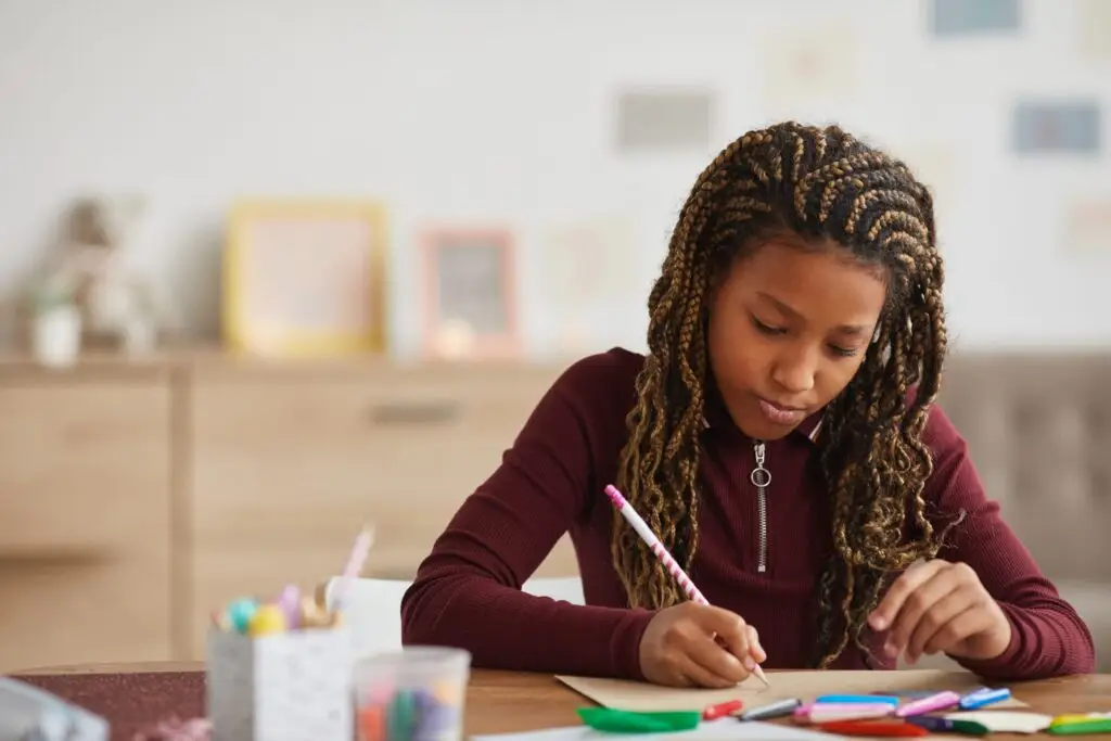 A young girl with braided hair, demonstrating fine motor skills, sits at a table focused on writing with a pencil. The table is scattered with colorful markers and pens. She wears a maroon top, and the background shows blurred frames on the wall.