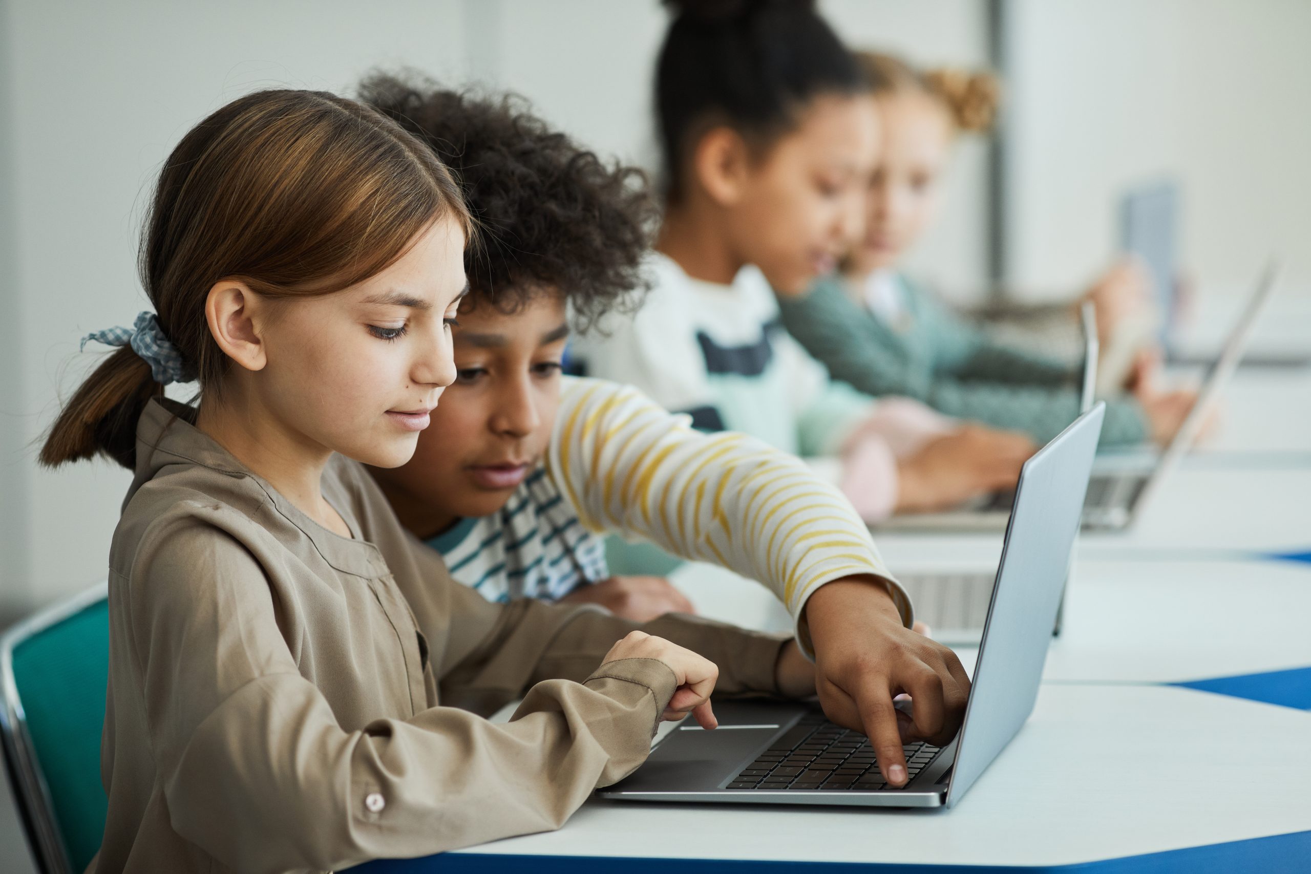 Children sitting in a row at a table using laptops, with two focused on the screen in the foreground. In this classroom setting, they embrace "Kids and Computer Safety: The 5 Internet Safety Rules" to ensure a secure online learning environment.