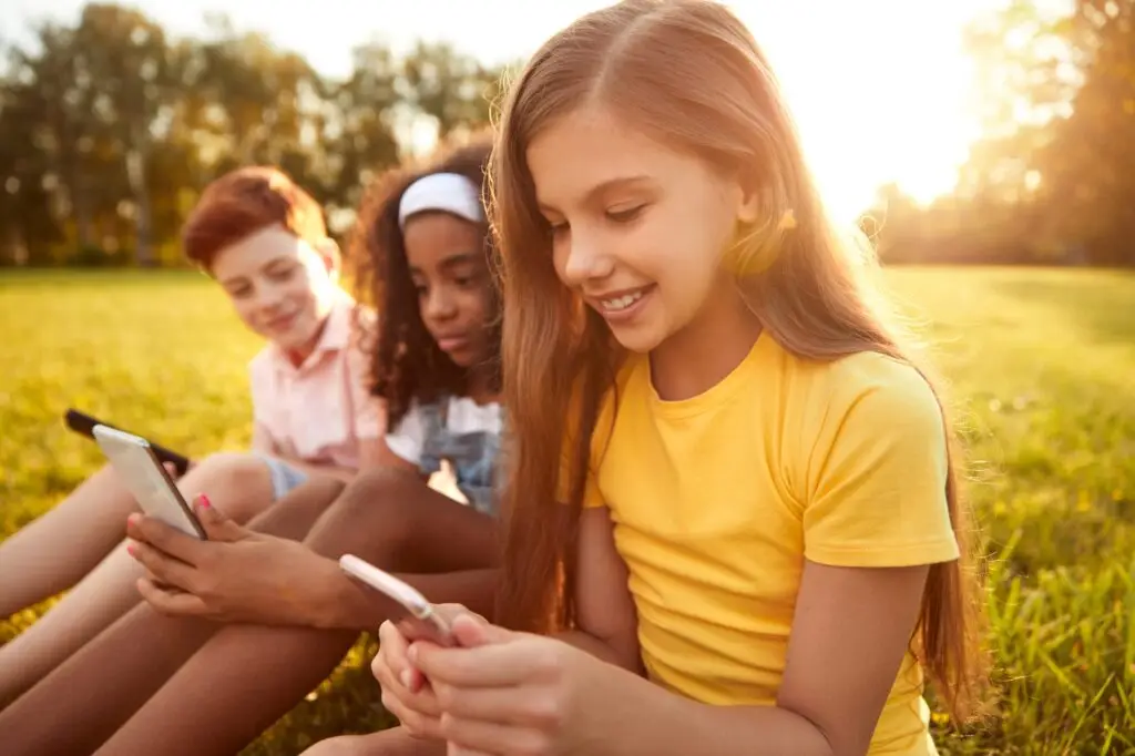 Three children sitting on grass in a park are using smartphones, raising the question: What age should a child get a cell phone? The girl in the foreground is smiling and wearing a yellow shirt. The other two kids are slightly blurred, enjoying the sunny day amidst trees and sunlight.