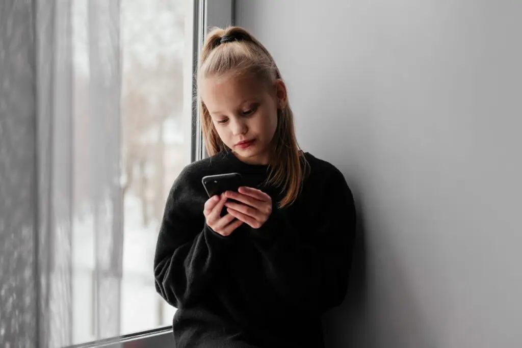 A young girl with long blonde hair tied in a high ponytail stands by a window, wearing a black sweater. She glances down at her smartphone, perhaps pondering how TikTok is affecting our kids. The background is soft and neutral, with the blurred view outside emphasizing her contemplation.