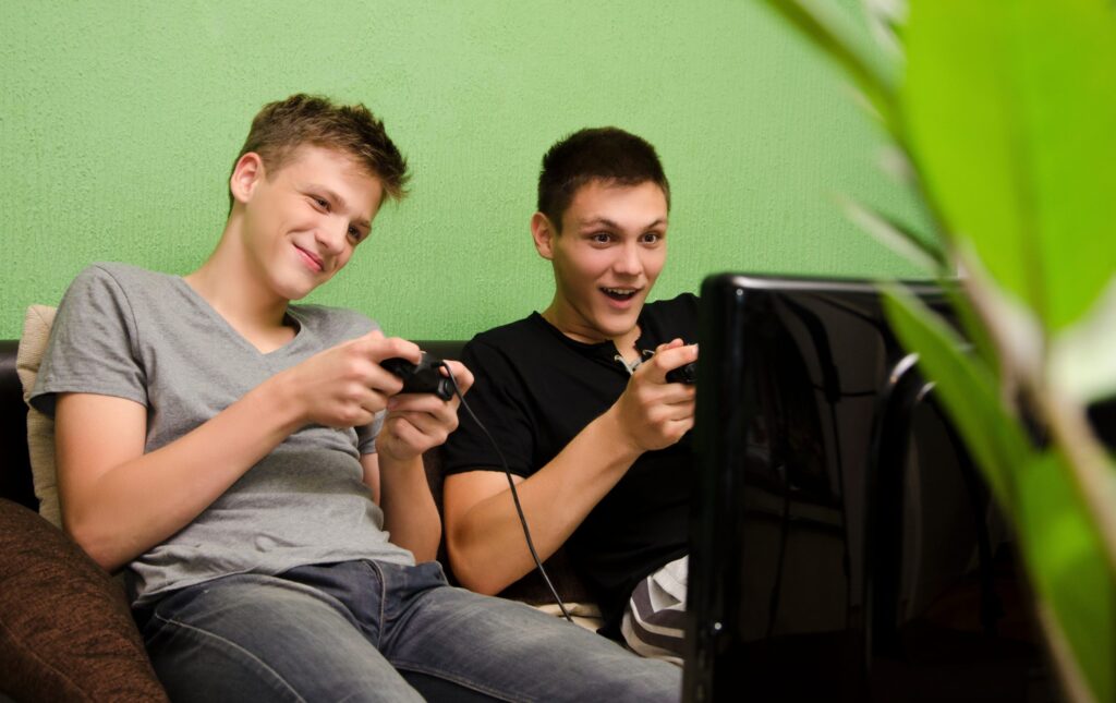 Two young men are sitting on a couch, gaming controllers in hand, looking excited and engaged as they play. In the foreground, the large TV screen mirrors their enthusiasm. A green wall serves as the backdrop, with a plant partially framing this scene of aspiring online streamers perfecting their Twitch skills.