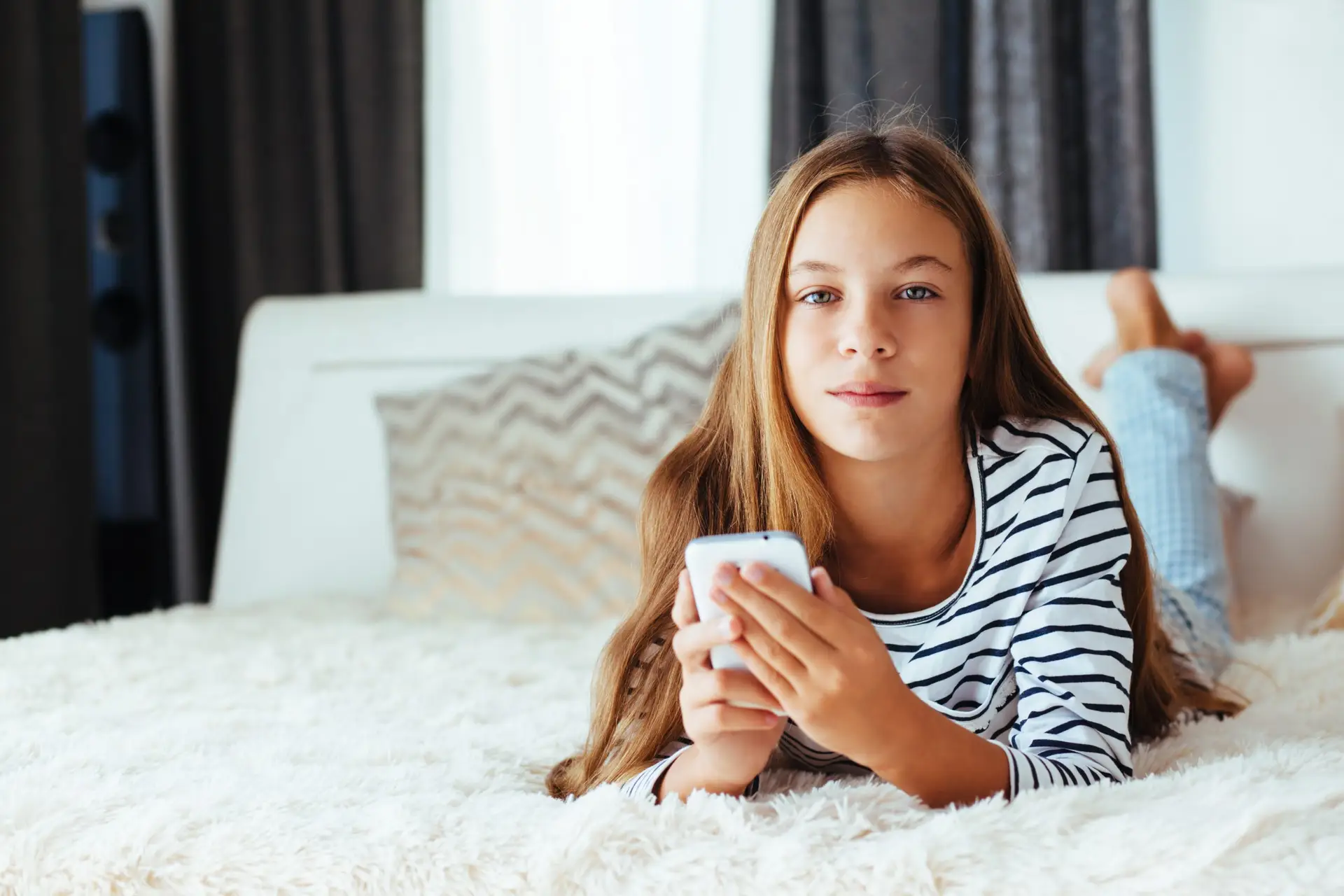 A young girl with long hair lies on a bed, cradling her smartphone like a secret treasure. She wears a striped shirt and gazes at the camera, curiosity in her eyes. The room is softly lit with neutral tones, capturing the essence of safe spaces—much like a Troomi phone's safelisting feature.