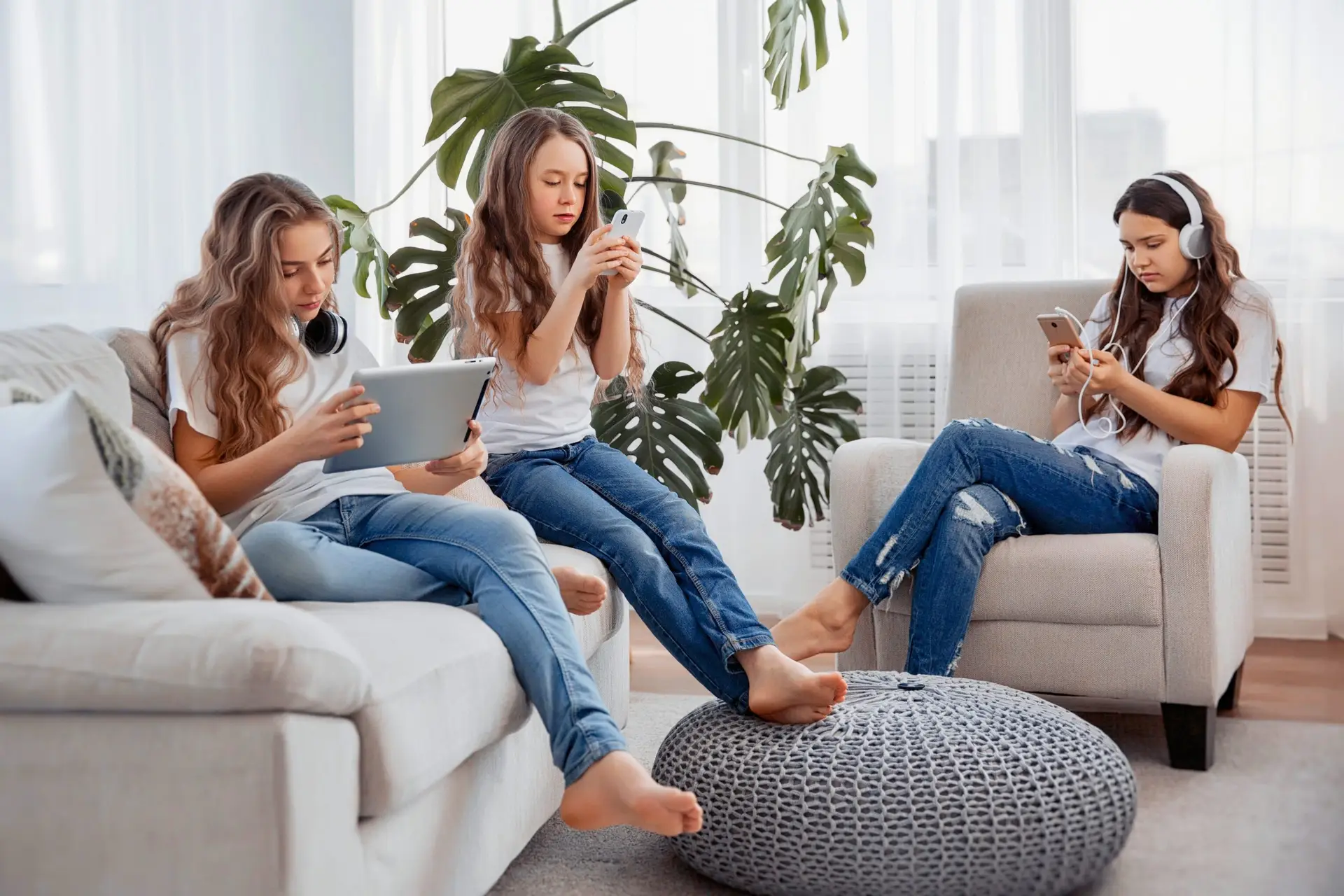 Three children sit in a bright room. One is on a couch with a tablet, another lounges in an armchair wearing headphones using a phone, and the last one sits on the floor engrossed in her cell phone. They are all focused on their devices, with a large plant quietly observing from the background.