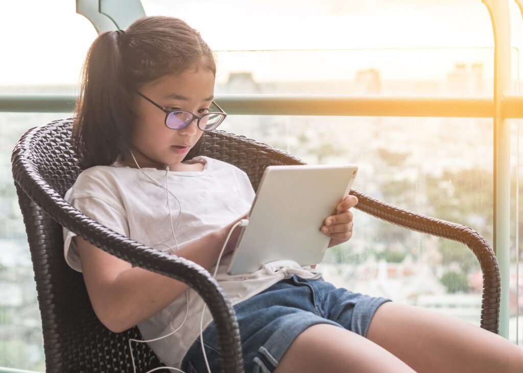 A young girl wearing glasses sits in a wicker chair, engrossed in her tablet as she explores Seven Ways to Practice Internet Safety for Kids. With earphones in, the sunlight filters through a glass wall behind her, creating a warm ambiance.