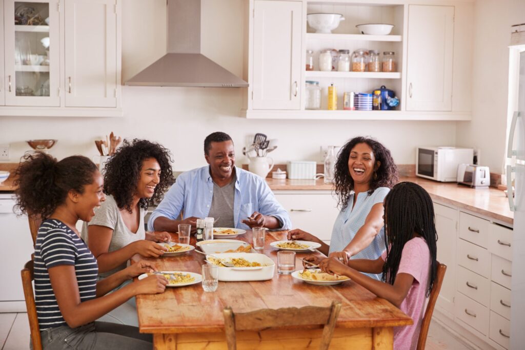 A family of five sits around a wooden table in a kitchen, enjoying a meal together. They are smiling and laughing, discussing 50 fresh dinner conversation topics for families, creating a warm, cozy atmosphere. Dishes of food are spread out on the table, and natural light fills the room.