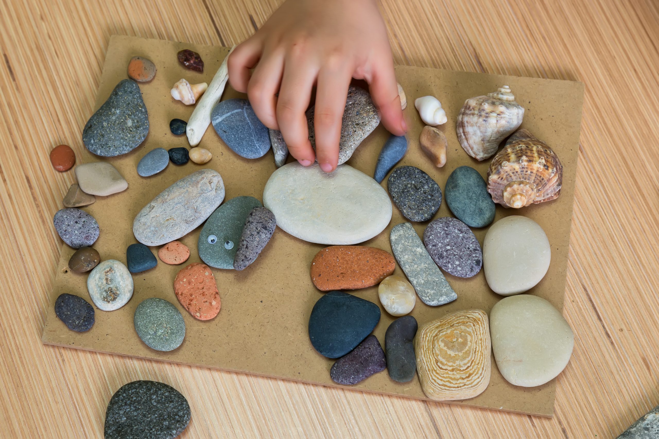 A child's hand arranges various colorful pebbles and shells on a wooden board, exploring the fascinating world of geology and minerals for kids. The surface is filled with stones of different shapes and textures, including elongated shells and one with googly eyes, all set against a light wood table.