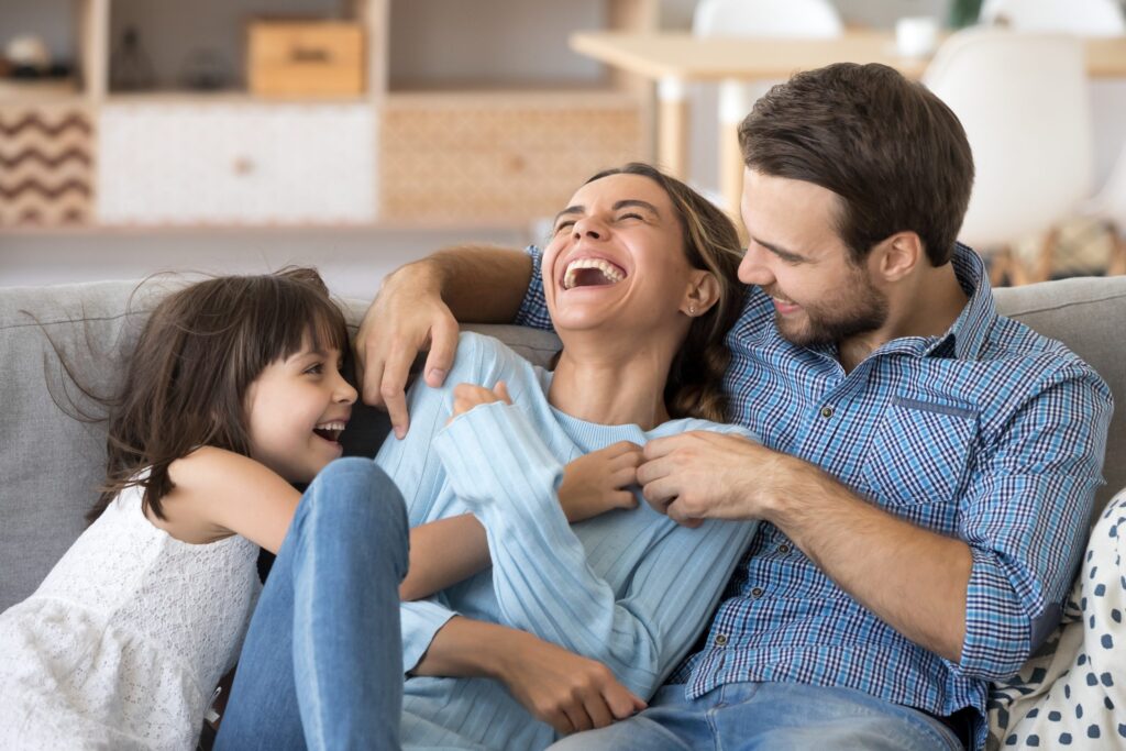 A joyful family of three, including a woman, man, and a young girl, laughing together while sitting on a couch in a bright living room. The woman is wearing a blue sweater and the man is in a checked shirt. The girl joyfully leans on them, embodying why you should take a break from social media.