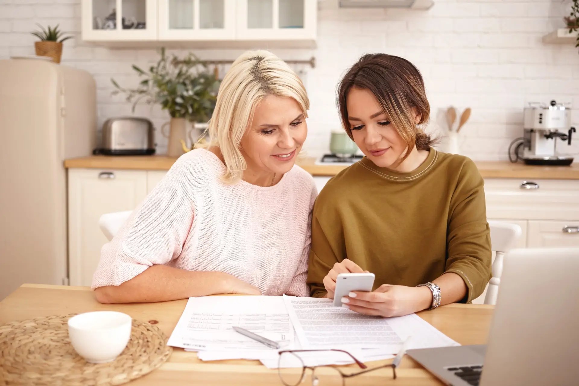 Two women sit at a kitchen table, engrossed in "What is Bitcoin: A Beginner's Guide to Cryptocurrencies" while reviewing documents and using a smartphone. Nearby, a laptop and coffee cup add to the scene. The bright kitchen features light-colored cabinets and a coffee maker in the background.