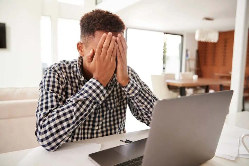 A person sits at a table with their head in their hands in front of a laptop, looking stressed or frustrated, as if pondering the question: "What Is Digital Self-Harm?" The room is bright with a modern dining area in the background.