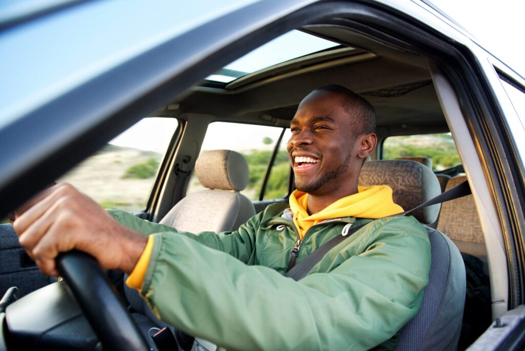 A person in a green jacket and yellow hoodie is happily driving a car, the window open to the sun's rays. The blurred landscape whizzes by, reminding us of the risks of distracted driving—Five Reasons Why You Shouldn’t Text and Drive. Safety ensures every journey stays joyful.