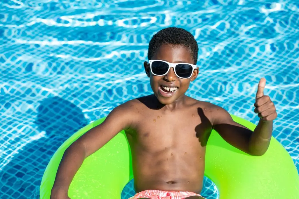 A child wearing white sunglasses is smiling and giving a thumbs-up, embodying the essence of summer safety for kids, as they sit on a bright green inflatable ring in a clear blue swimming pool. The refreshing scene captures the joy and care of sunny days by the water.