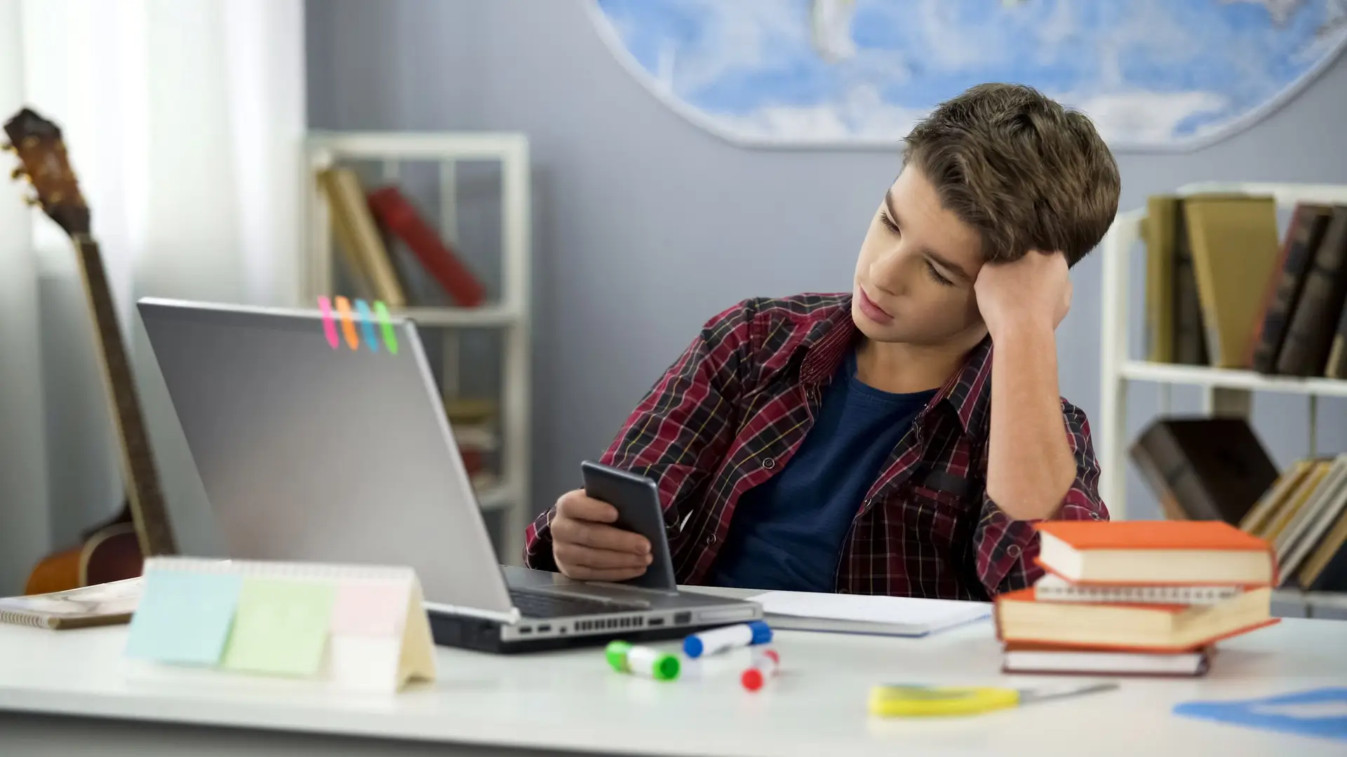 A teenage boy in a plaid shirt sits at a desk, studying the question, "What Causes Procrastination and How Can We Stop It?" He looks thoughtfully at his phone. His desk is cluttered with books, papers, and colored markers. A guitar stands nearby, while a world map adorns the wall.