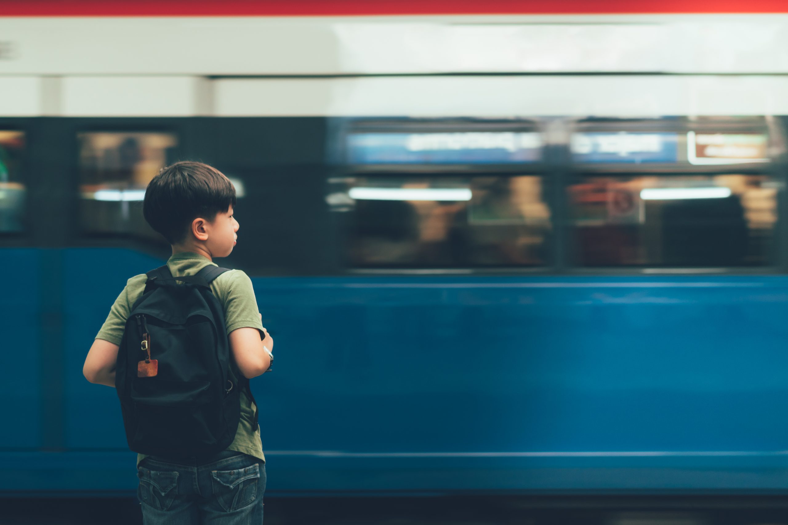 A young boy with a backpack stands still on a train platform, observing the blue and white train speed by in a blurred motion. Focused and attentive amid the bustling scene, he embodies one of 3 ways to teach kids about public transport systems: real-world exploration.