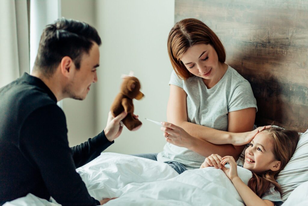 A family scene unfolds with a man playfully holding a teddy bear, a woman nearby reading prescription drug facts on a thermometer, and a young girl lying in bed smiling. The woman's gentle touch on the girl's head creates a warm and caring atmosphere.