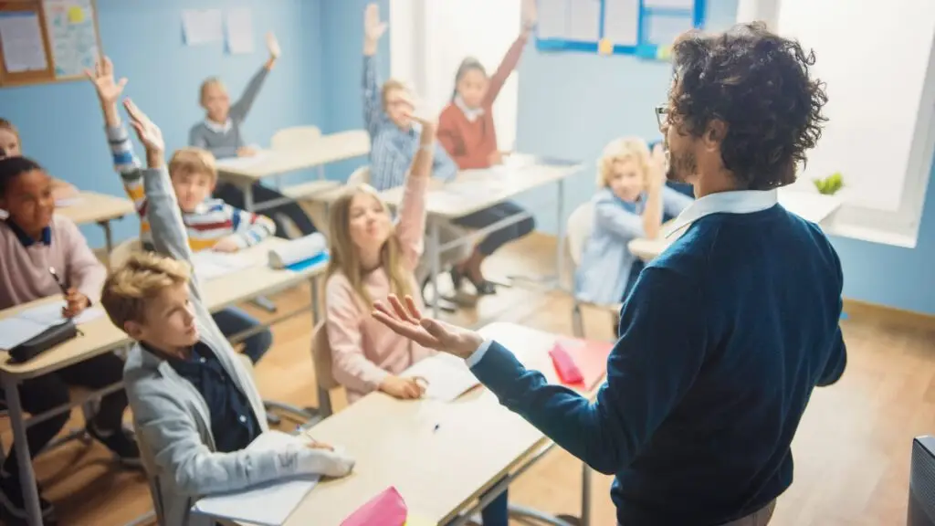 A teacher stands at the front of a classroom, engaging with students seated at desks. Several students raise their hands eagerly, ready to discuss the Four Rules of Active Listening for Kids. The room is bright and organized, adorned with educational materials on the walls.