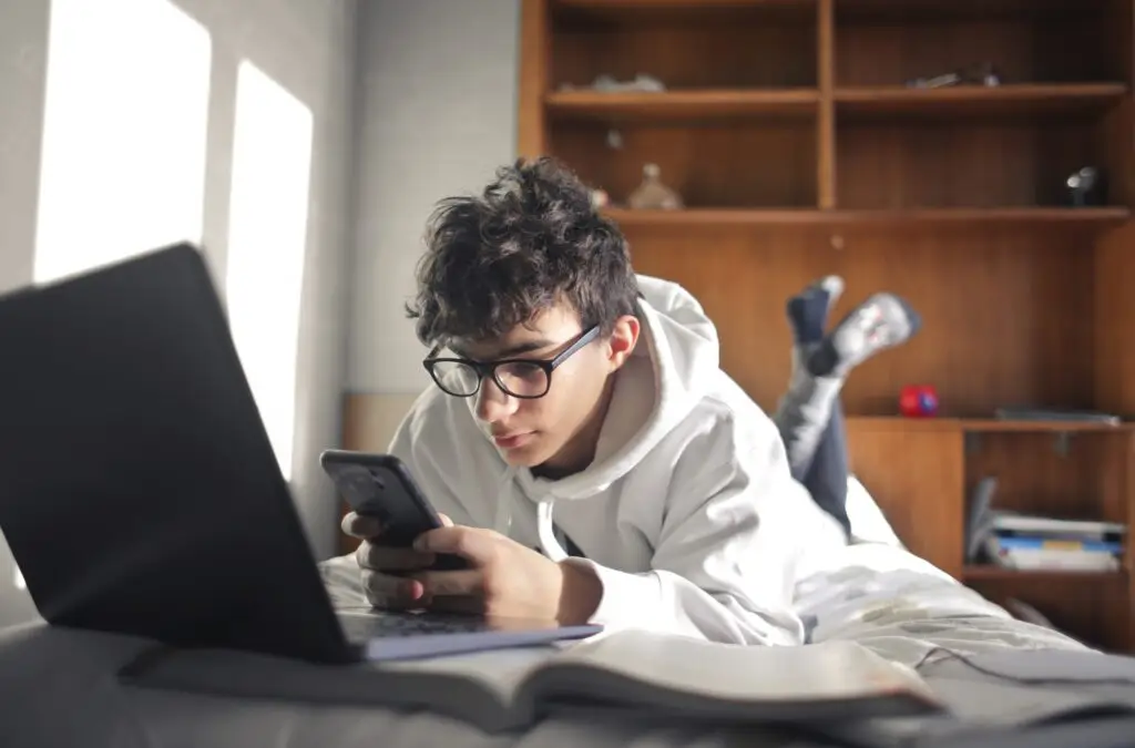 A young person wearing glasses and a white hoodie is lying on their stomach on a bed, scrolling through "Is TikTok Safe For Kids? The Ultimate Guide For Parents" on their smartphone. An open laptop and a book are nearby, with wooden shelves visible in the background.