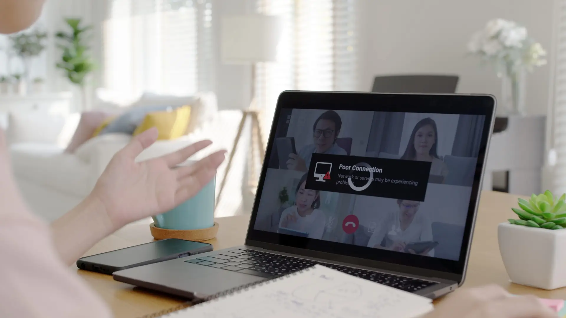 A person engages in a video conference on a laptop at home, encountering a poor connection message on the screen. Gesturing with one hand while contemplating troubleshooting steps, they sit in a softly lit room with a notebook and smartphone resting on the table, creating a cozy setup.