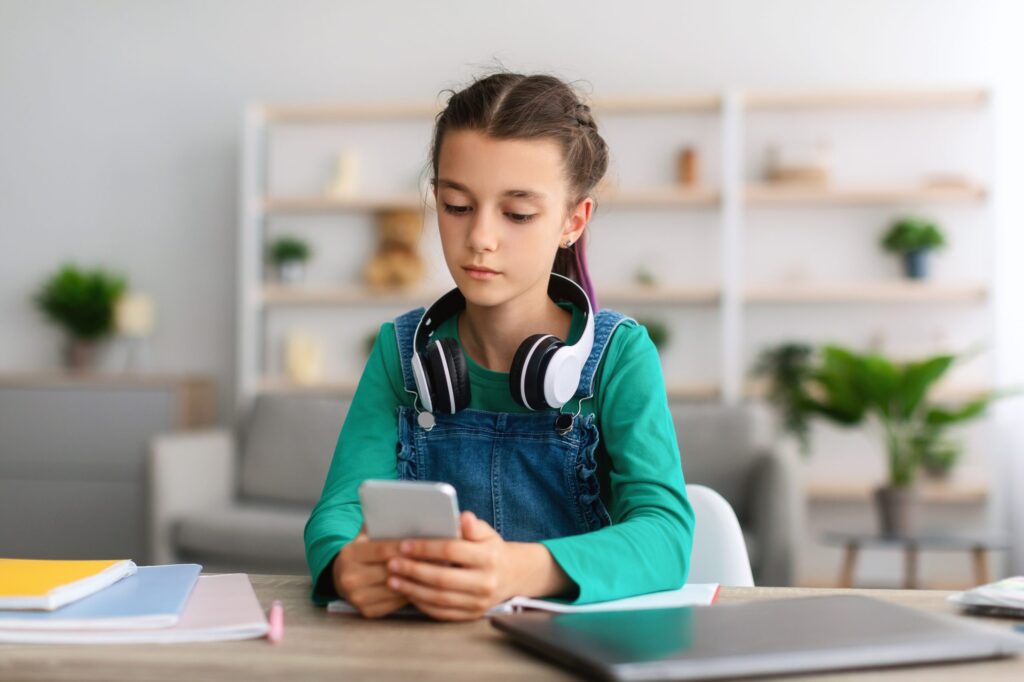 A young girl with headphones resting around her neck gazes at a smartphone while seated at a table with notebooks and a laptop. In a room filled with shelves and plants, she explores how Troomi can help address why cell phones might be bad for kids.