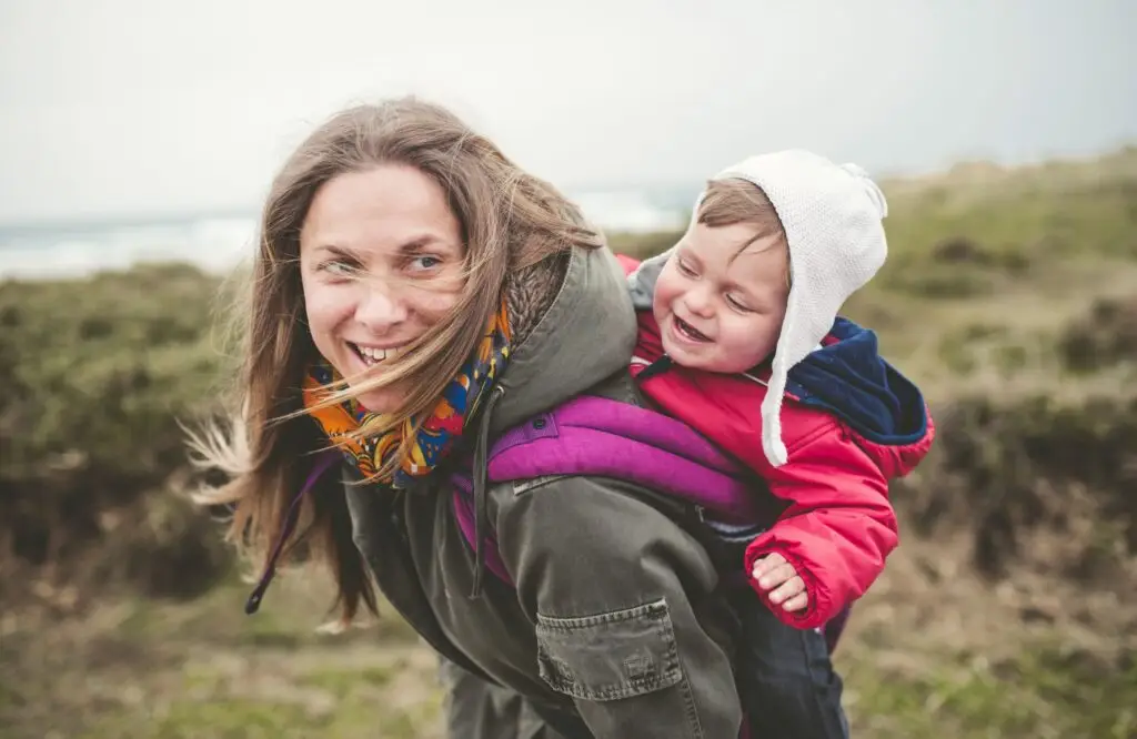 A smiling woman carries a happy toddler on her back, dressed in a red jacket and white hat. They're outdoors in a grassy area with the sea behind them, enjoying a playful, windy day—one of many reasons you should start hiking with your baby.