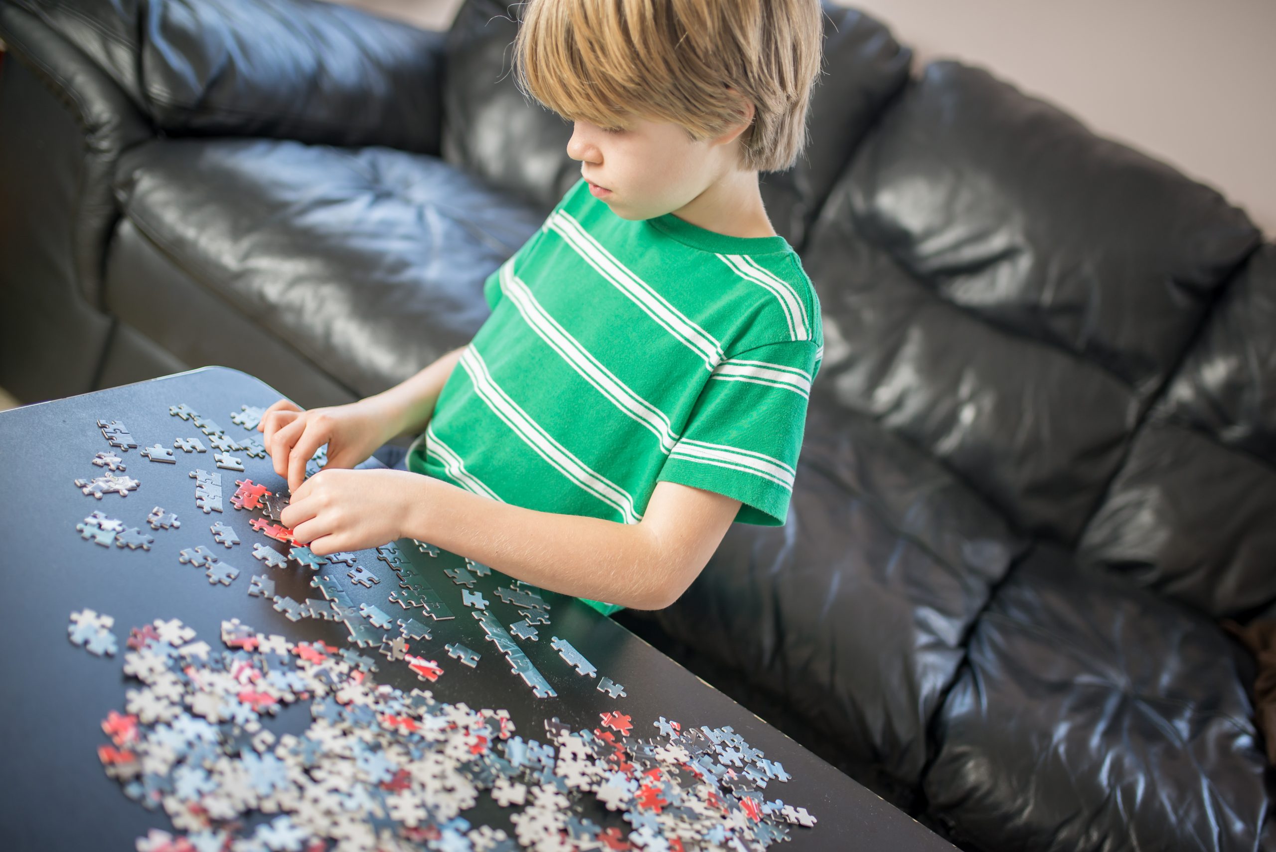 A young child in a green striped shirt is focused on assembling a jigsaw puzzle at a table, reminiscent of the benefits of old-fashioned wooden toys. A black leather sofa sits quietly in the background.