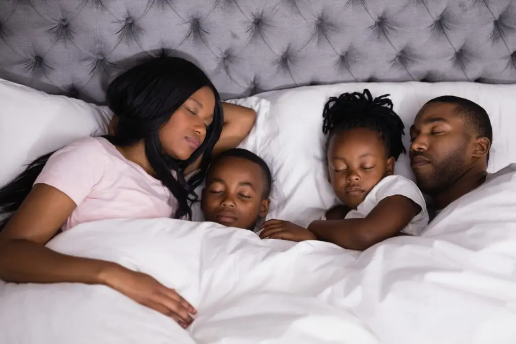 A family of four sleeps peacefully in a bed, each following their own sleep as needed routine. The parents lie on either side of two children, all covered with a white blanket. The tufted gray headboard adds to the cozy and serene atmosphere.