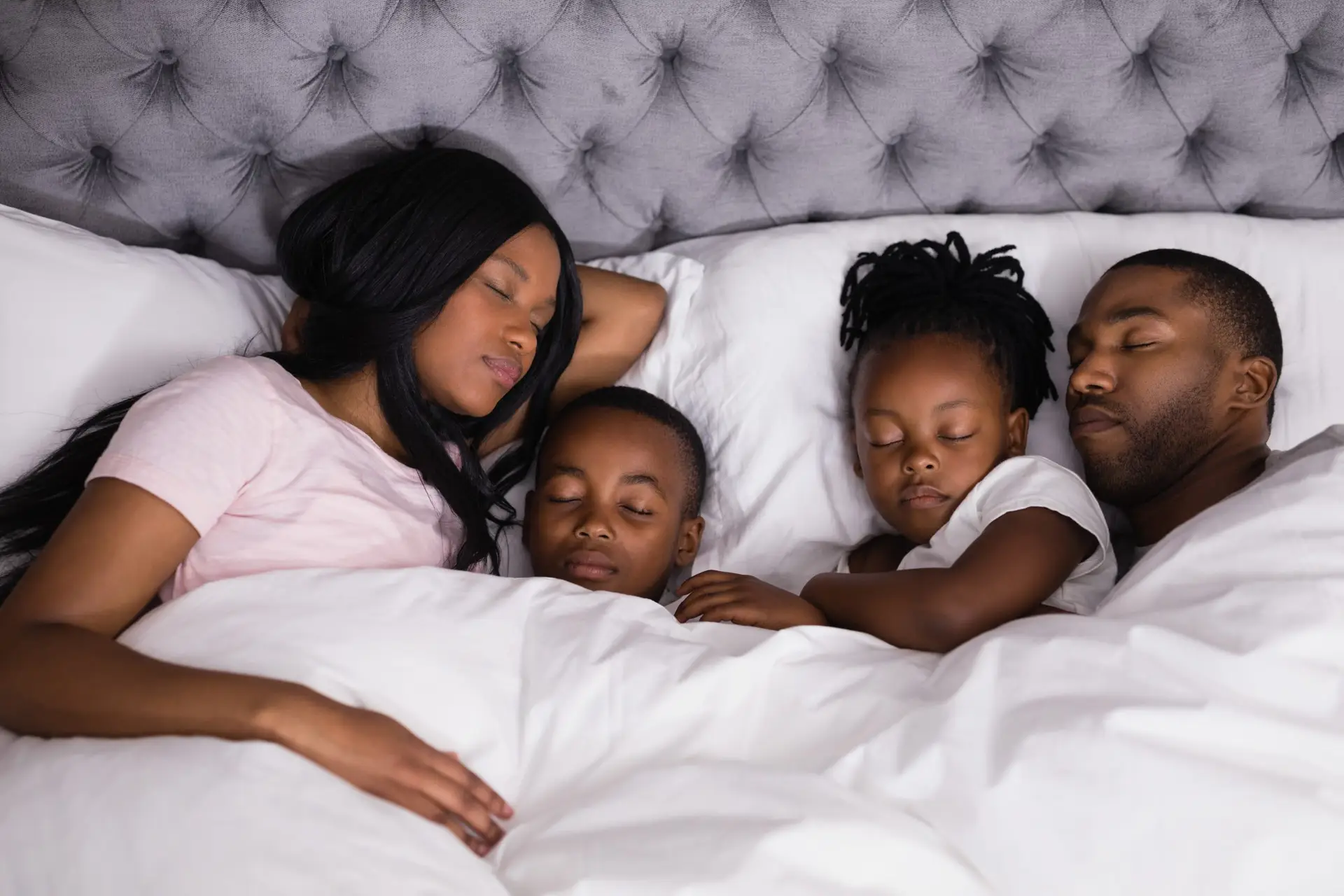 A family of four sleeps peacefully in a bed, each following their own sleep as needed routine. The parents lie on either side of two children, all covered with a white blanket. The tufted gray headboard adds to the cozy and serene atmosphere.