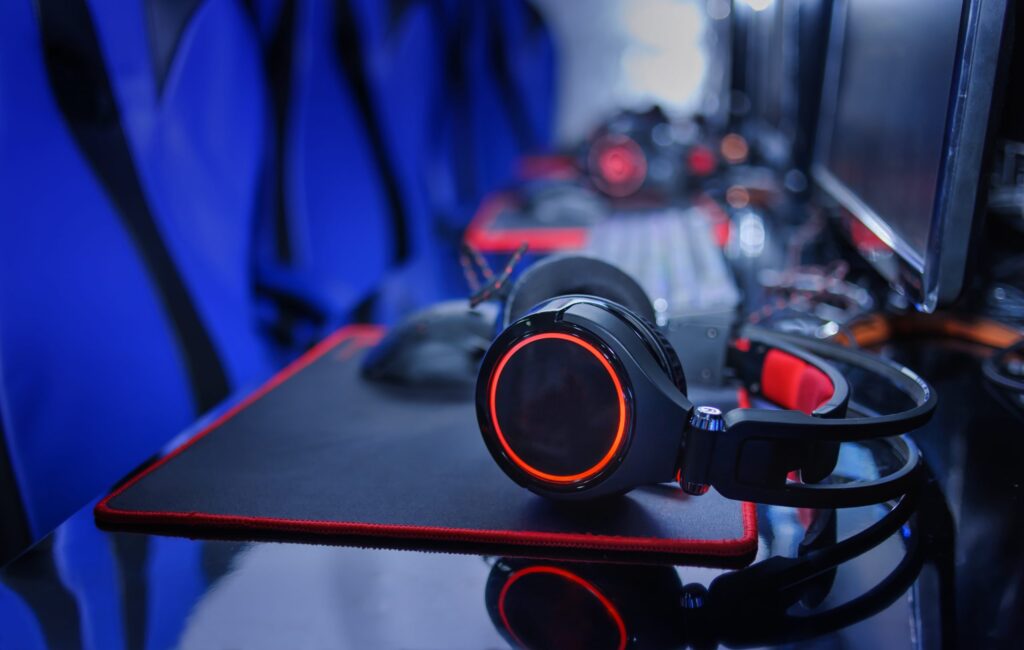 A row of gaming stations, perfect for diving into the world of esports tournaments, features sleek black headphones with red LED lighting resting on a black and red mousepad. Blue gaming chairs and computer screens in the background complete this immersive gaming setup.