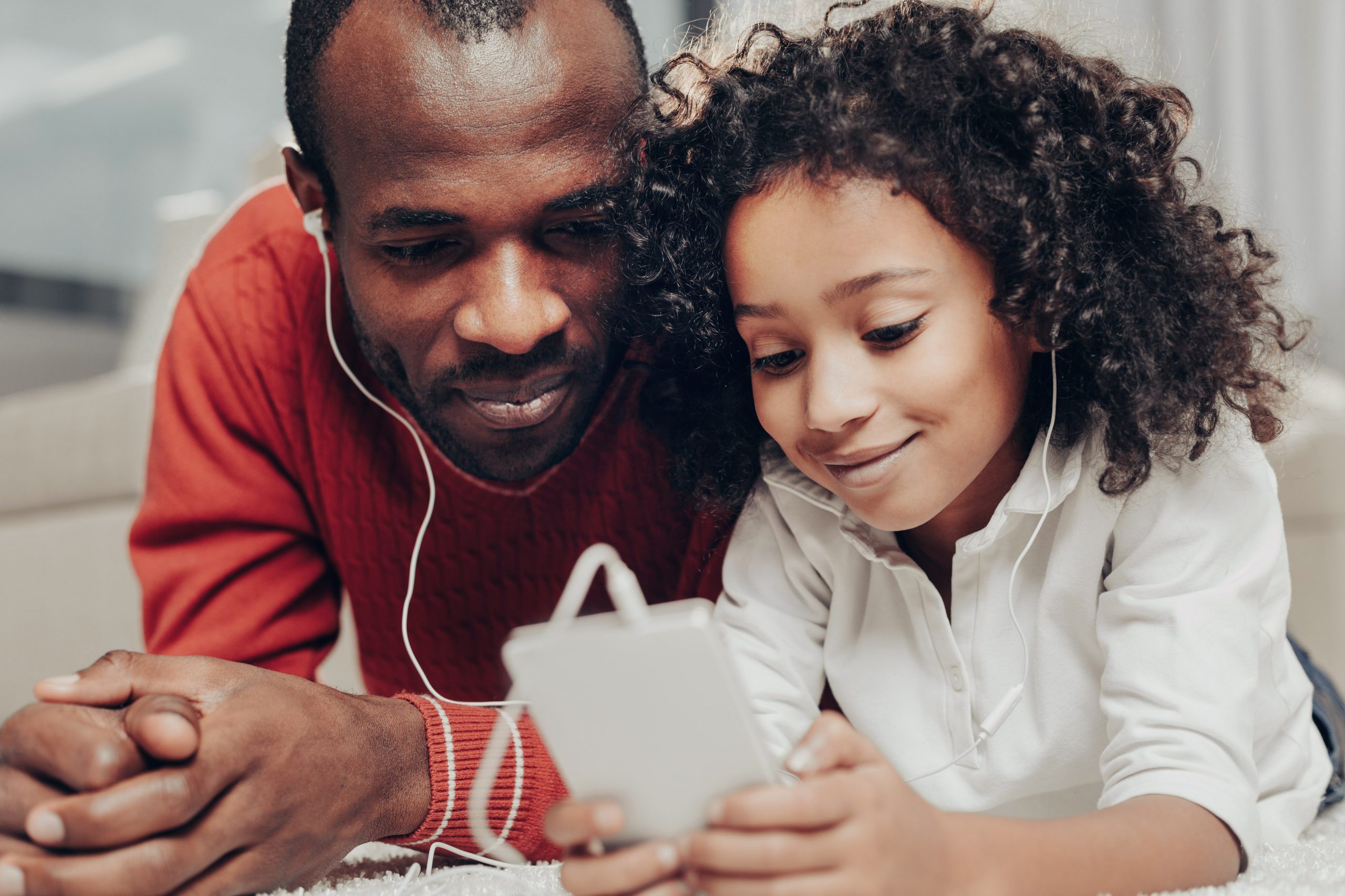 A man and a child lie on their stomachs, sharing earphones while using the best app for teaching music online. The man wears a red sweater, and the child is in a white shirt. They appear engaged and happy, focused on the enriching content on the smartphone screen.