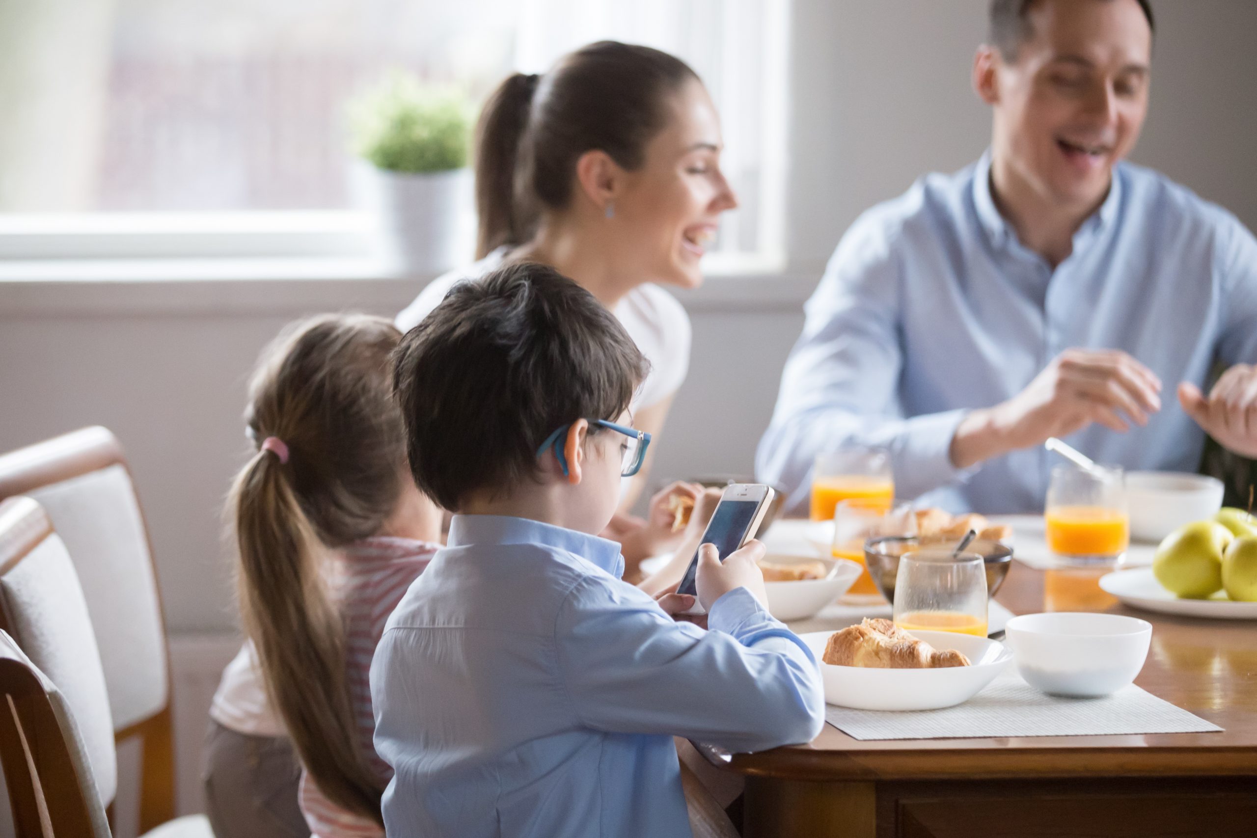 A family of four sits at a dining table having breakfast. An adult man and woman are smiling and talking, while a boy in glasses looks at a smartphone, perhaps learning about cell phone etiquette rules your child should know. A girl with long hair is seated next to the boy. The table has croissants, fruit, and juice.