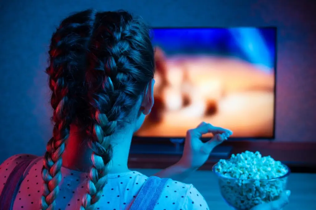 A person with braided hair is sitting in front of a TV screen featuring scenic views, possibly indulging in one of the Six Movies and TV Shows About Teen Addiction, while holding a bowl of popcorn. The room is dimly lit, reflecting blue and red hues.