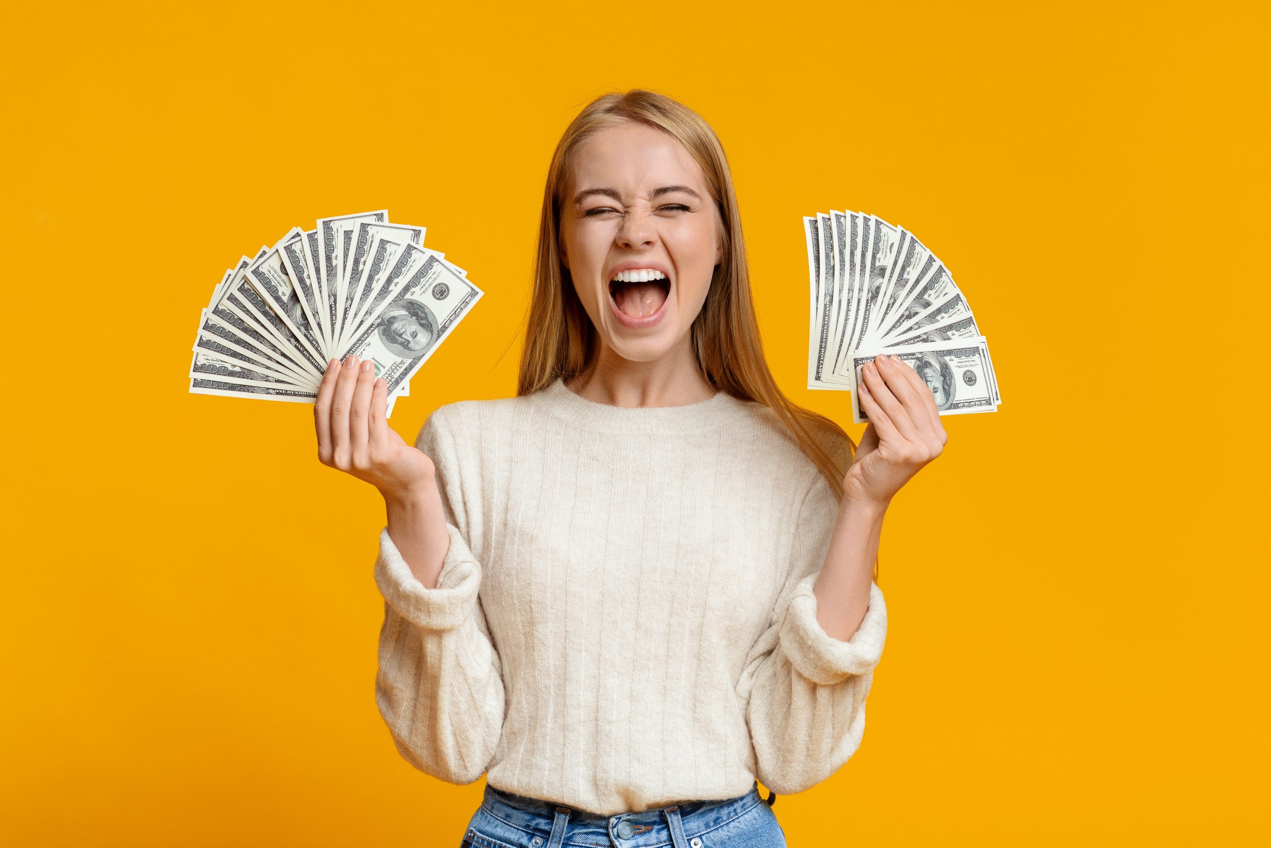 A woman stands against a bright yellow background, smiling broadly and holding a fan of U.S. dollar bills in each hand—a perfect representation of mastering money management. She wears a light-colored sweater and jeans, embodying the savvy spirit found in "Ten Simple Tips for Teens Managing Money.