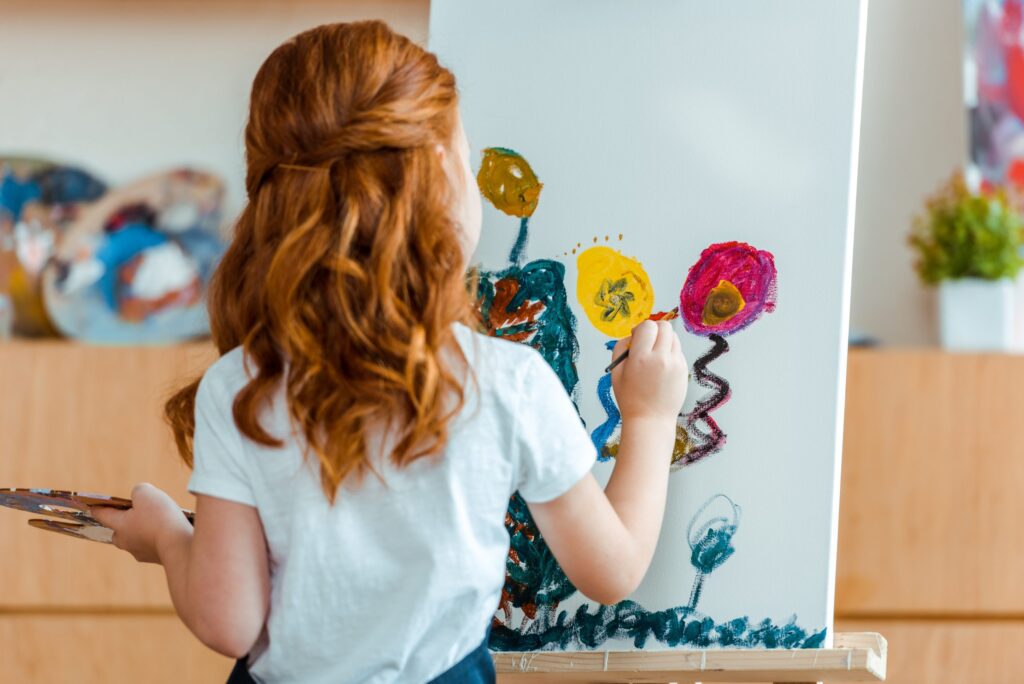 A young girl with red hair paints colorful flowers on a canvas, using her creative skills nurtured by child development programs. She holds a palette in one hand and brushes with the other. The room is softly lit, and plants are visible in the background.