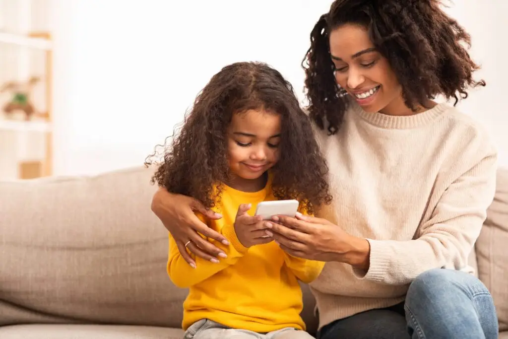 A woman and a child with curly hair sit on a couch, the child in a yellow sweater, engrossed in a smartphone as the woman, dressed in cream, smiles at the screen. They appear to be enjoying their time together while exploring how to monitor text messages for free.