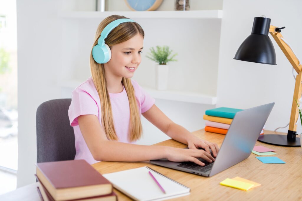 A girl wearing blue headphones sits at a wooden desk, using a laptop to explore how to create Gmail emails for kids under 13. She is smiling in her pink shirt, surrounded by books, colorful sticky notes, and a black desk lamp, with a small plant adding a touch of green in the background.