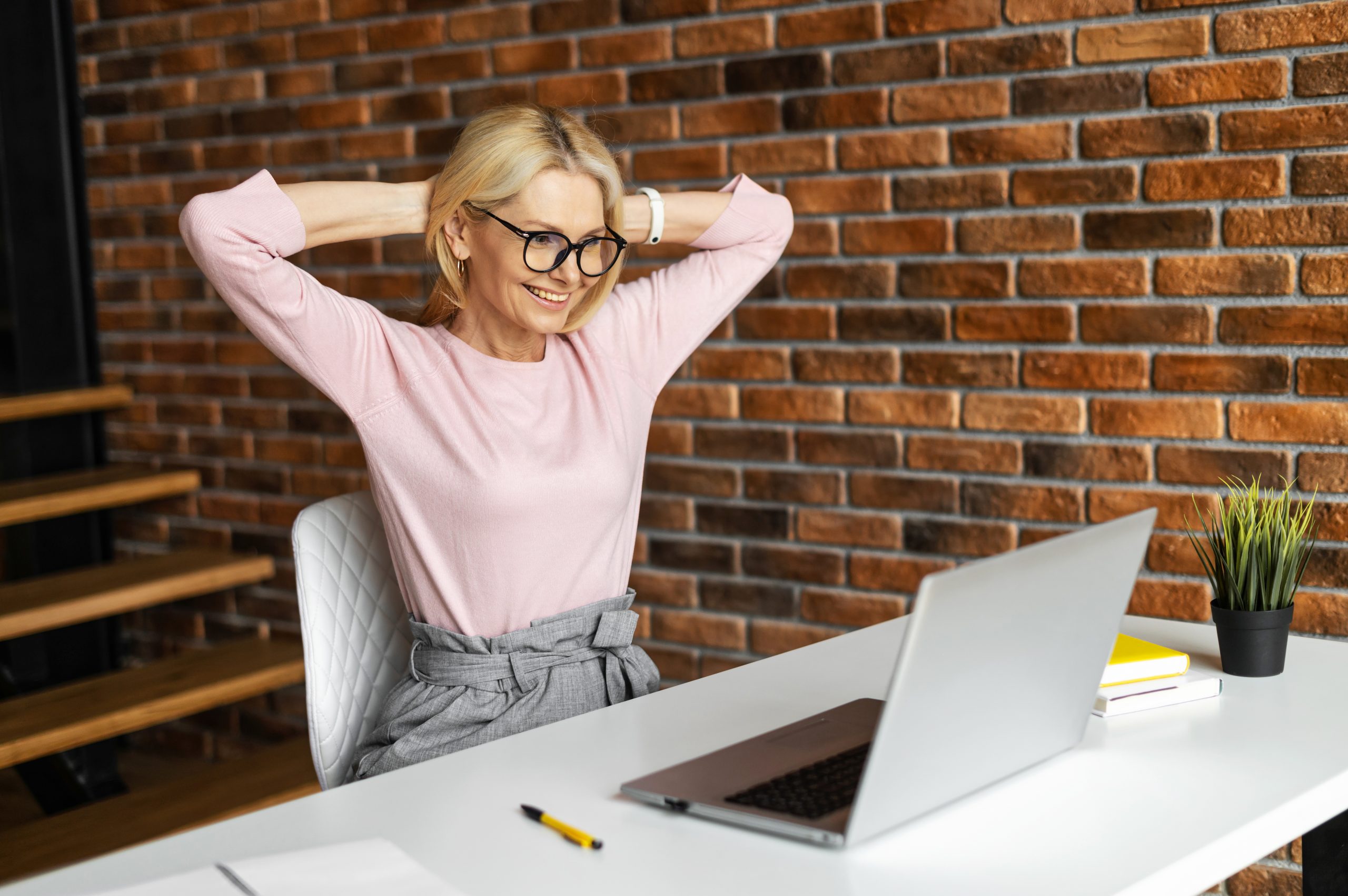 A woman with glasses and blonde hair sits at a desk, smiling at a laptop. Her pink shirt and gray pants enhance her confident aura as she stands up straight. Against the backdrop of a brick wall, a notebook, pen, and small plant keep her company while she enjoys the benefits of good posture.