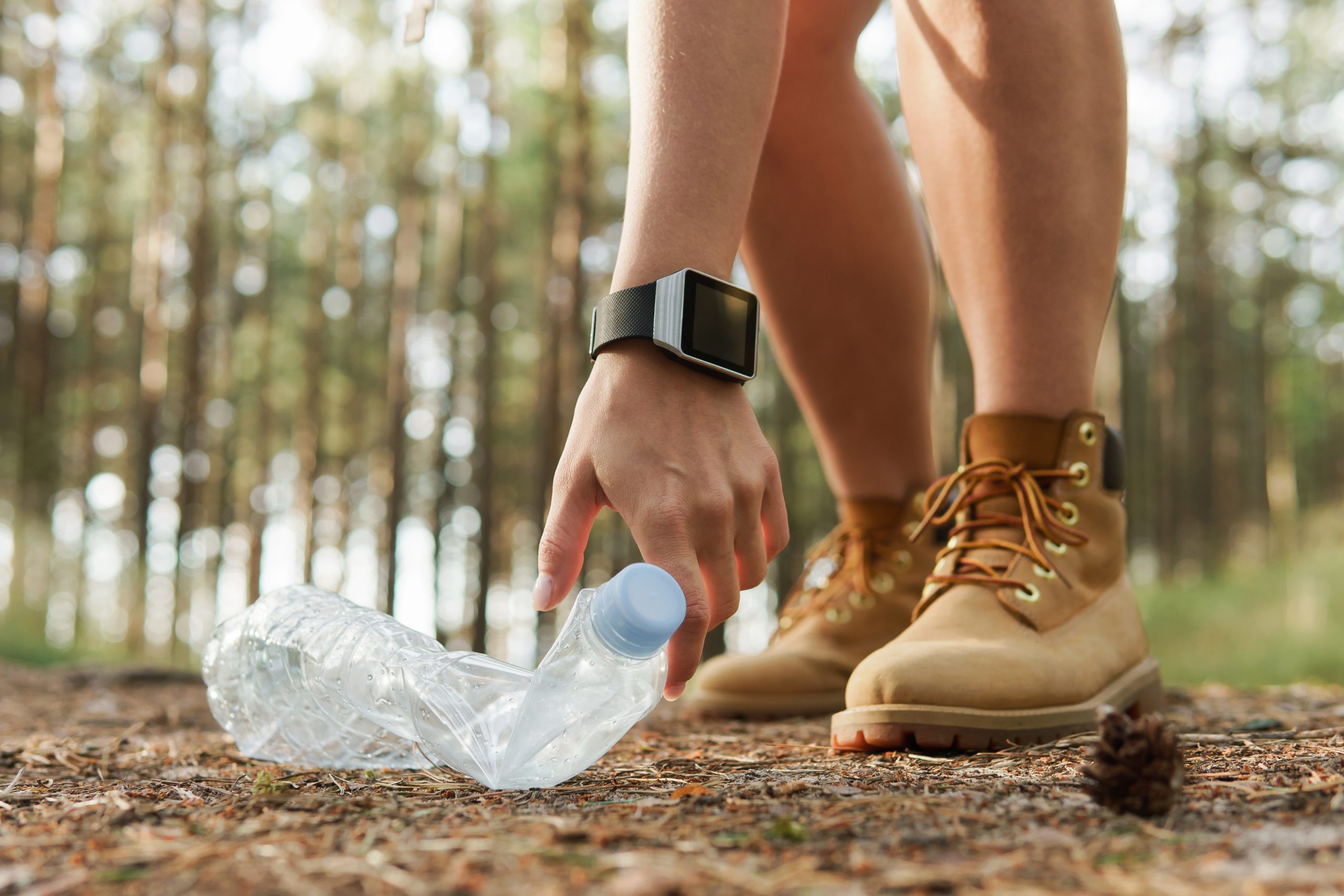 A person wearing brown boots and a smartwatch demonstrates one of the Five Ways to Maintain Clean Trails When Hiking by thoughtfully picking up a crumpled plastic bottle from the forest floor. The lush trees and natural surroundings stand majestically in the background.