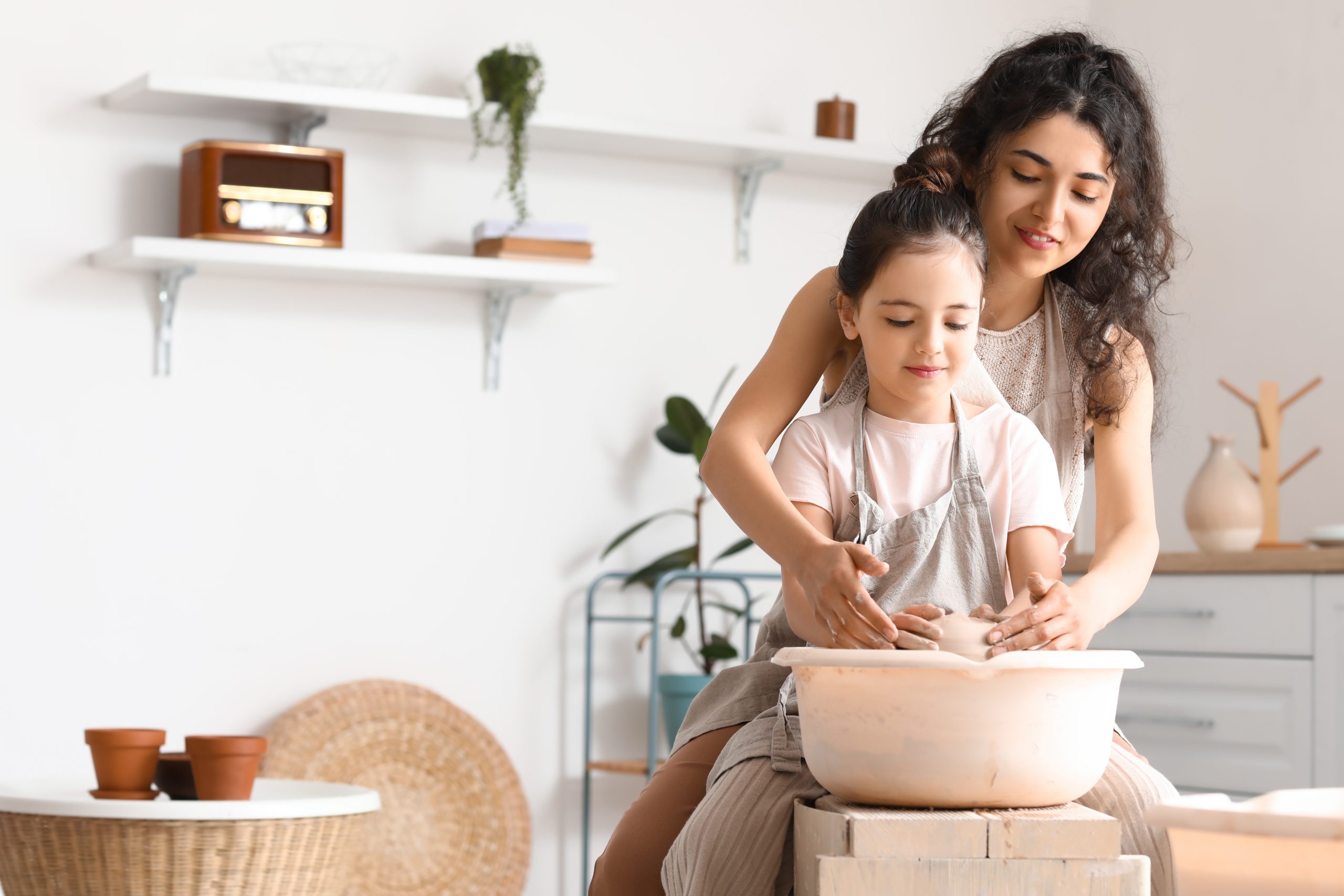 A woman and a young girl, embodying one of the five reasons to practice pottery for kids, are sculpting clay on a pottery wheel in a bright room with plants and shelves. Their smiles reflect their focus and joy in this creative activity.