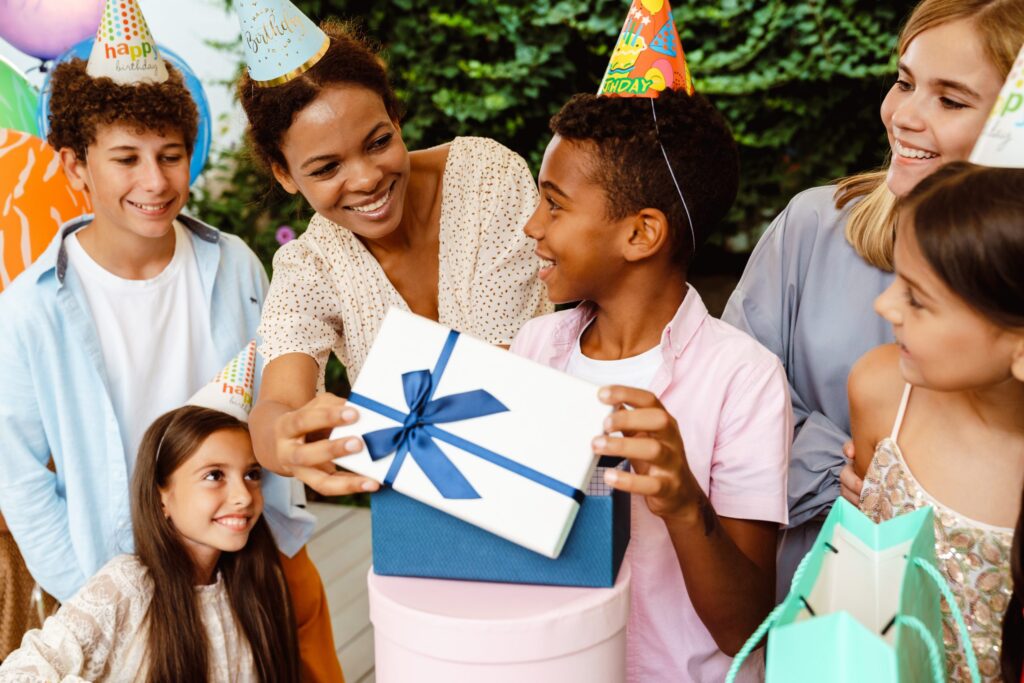 A group of children and an adult are enjoying a budget-friendly birthday party. They smile under colorful hats, with one child proudly holding a wrapped gift. Balloons surrounding the greenery backdrop make for delightful celebration ideas that bring joy without breaking the bank.