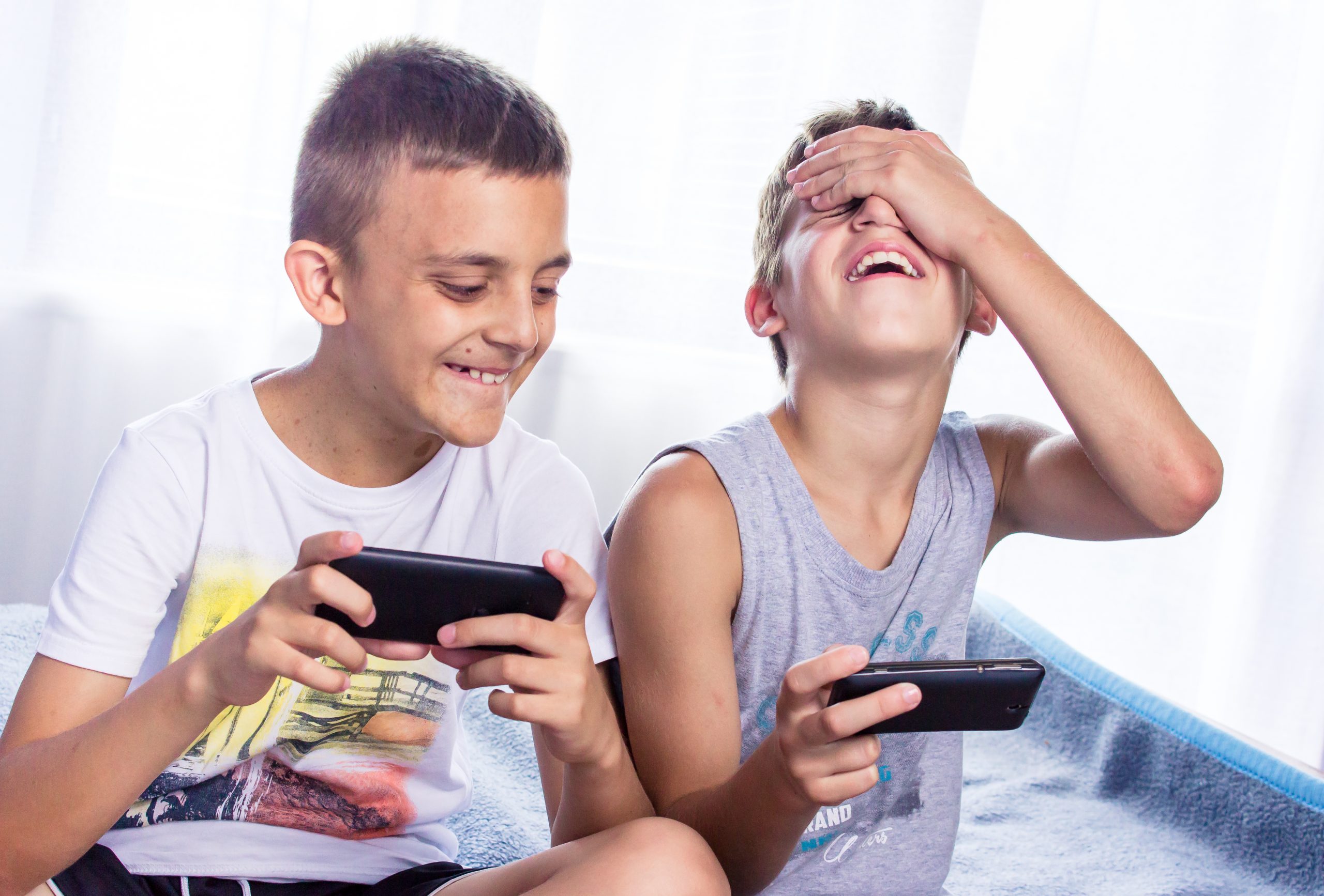 Two boys are sitting side by side, each holding a phone. The boy on the left is smiling while the boy on the right is laughing with his hand over his eyes. They appear to be enjoying a round of "Among Us" together, engrossed in its playful chaos. Both are wearing casual summer clothes.