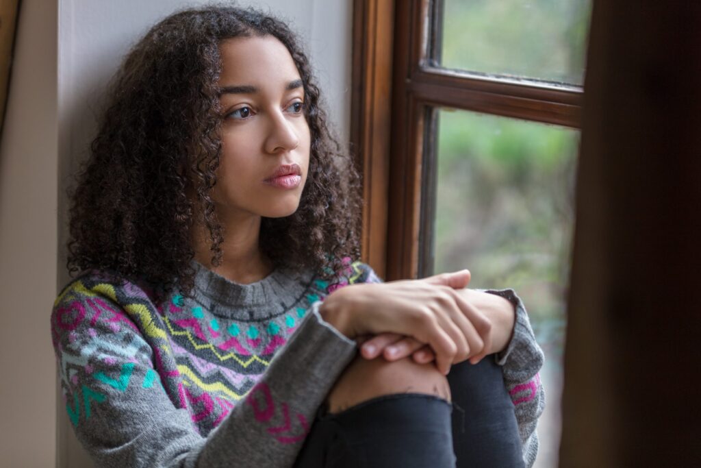 A person with curly hair sits by a window, wrapped in a patterned sweater, contemplating what to do when your kid isn’t invited. With arms around their knees, they gaze outside thoughtfully, as the room exudes a calm and introspective atmosphere.