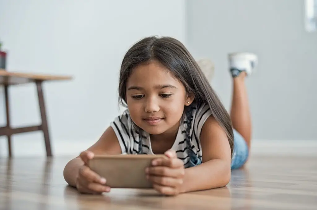 A young girl with long hair lies on her stomach on a wooden floor, engrossed in a language learning app for kids on her smartphone. She wears a striped shirt and denim shorts, focused entirely on the screen. In the background, a table with a plant adds a touch of greenery to the scene.