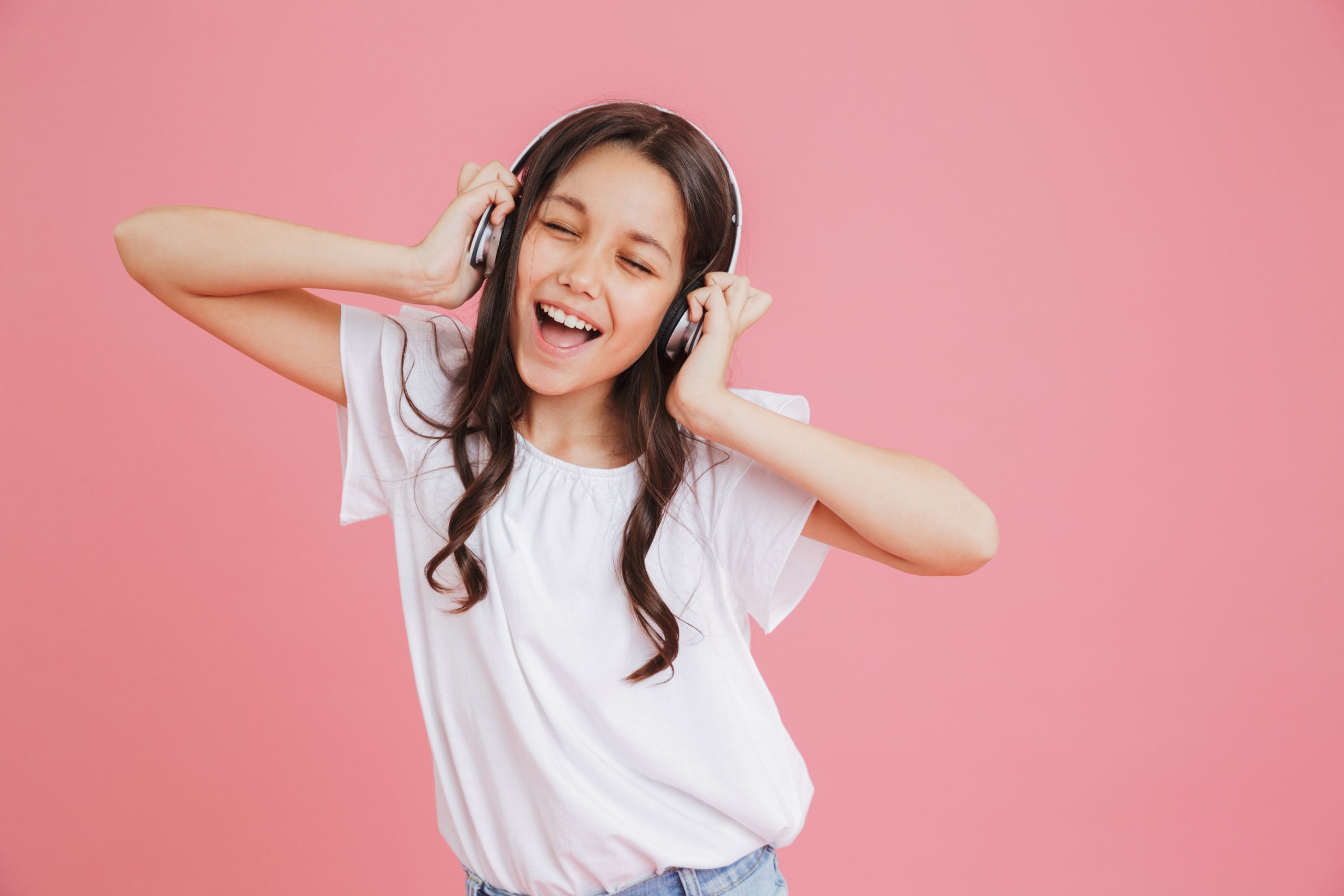 A young girl with long hair is wearing headphones, immersed in the joy of music from Spotify Kids. She smiles blissfully, her eyes closed, dressed in a white shirt against a pink background, completely captivated by her musical adventure.