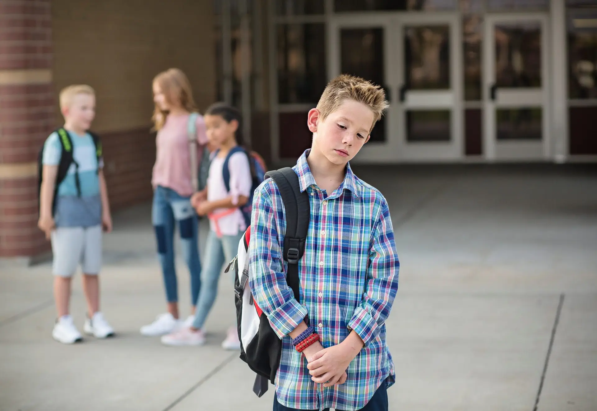 A boy with a backpack stands alone in the foreground, looking down with a somber expression, embodying how to deal with FOMO. In the background, slightly blurred, three other children chat near a school entrance. It's crucial for parents to learn ways to help bridged this gap.