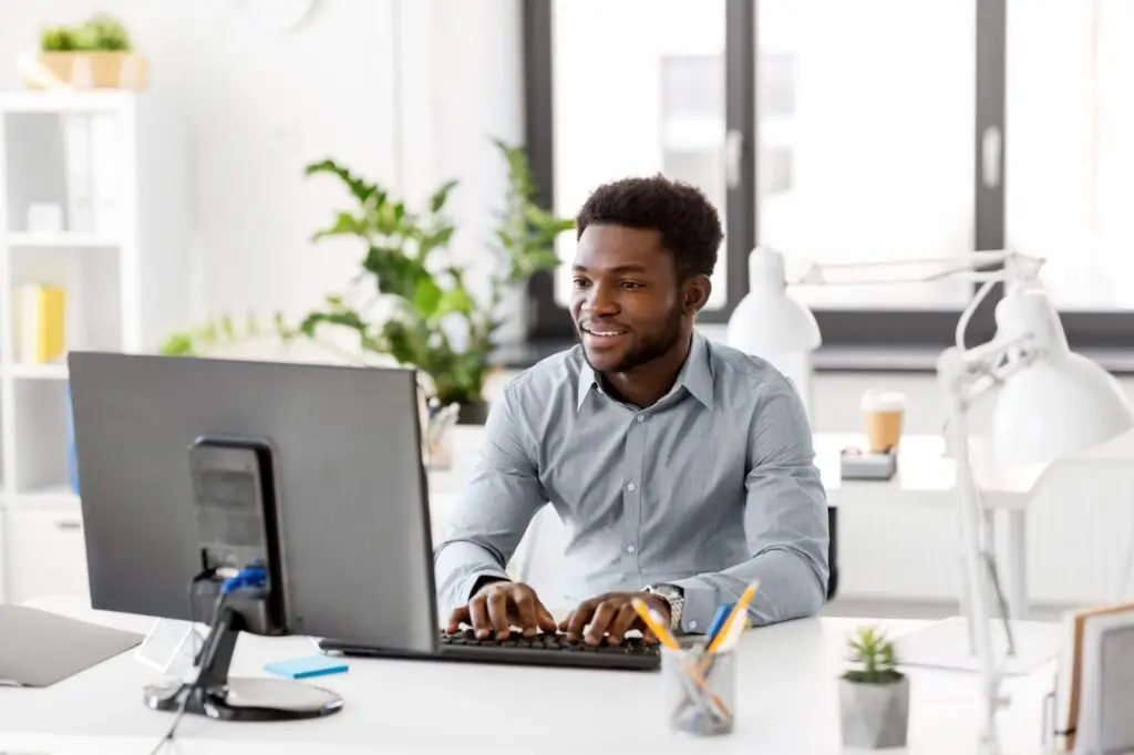 A man in a gray shirt is smiling while typing on a keyboard at his desk, curious about "What's the Deal with Web Push Notifications?" The organized desk boasts various office supplies and a small plant. The room is bright, with large windows and more plants in the background.